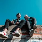 Two young people sitting on a stone wall under a vibrant blue sky, enjoying a sunny day together.